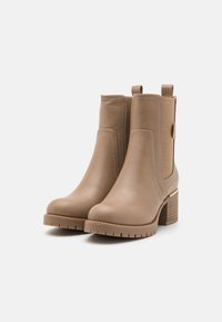 Bottines beiges avec une tige en cuir lisse, des panneaux latéraux élastiques et un talon bloc texturé. Présente une couture contrastée et un accent doré.