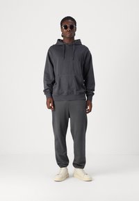 Grau-meliertes Baumwoll-Hoodie mit Fronttasche, kombiniert mit passenden Jogginghosen. Das Modell trägt Sonnenbrillen und cremefarbene Sneakers. Schlicht und entspannt geschnitten.