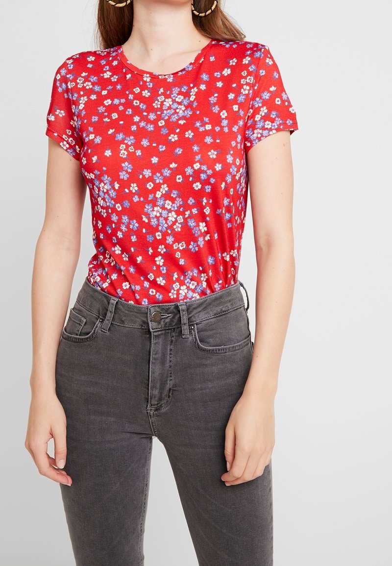 Camiseta roja de manga corta con pequeños patrones florales azules y blancos, combinada con unos vaqueros grises de cintura alta. Textura suave, escote redondo.