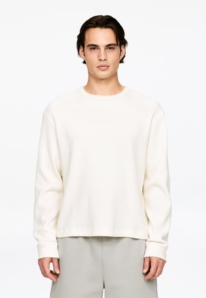 Light-colored waffle-knit sweatshirt with a round neckline and long sleeves, featuring cuffs and a relaxed fit, paired with light gray bottoms.