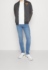 Mörkgrå zip-up Nike-jacka med orange detaljer, vit t-shirt under, ljusblå jeans och vita sneakers med svarta detaljer.