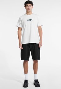 Tricou alb din bumbac cu logo-ul albastru "Guess Jeans", asortat cu pantaloni scurți din denim negri și pantofi negri cu șireturi, pe un fundal simplu.