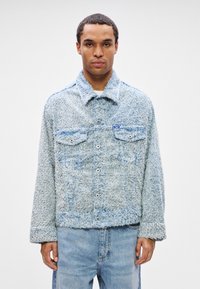 Man wearing light blue textured button-up jacket with chest pockets and light blue jeans, standing against plain white background.