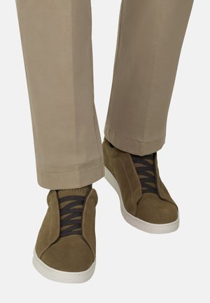 Braune Wildleder-Sneaker mit schwarzen Schnürsenkeln, kombiniert mit beigen Hosen mit geradem Bein, abgebildet von der Mitte der Wade abwärts vor weißem Hintergrund.