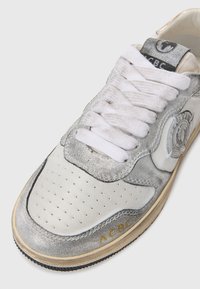 Weiße Leder-Sneaker mit silbernen Akzenten, durchbrochener Zehenpartie und strukturiertem Zunge. Ausgestattet mit weißen Schnürsenkeln und einem goldenen Logo an der Seite.