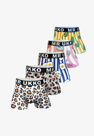 Hombre sonriente sin camiseta, con cabello rubio, de pie descalzo, vistiendo calzoncillos tipo bóxer blancos con estampado de leopardo en negro y marrón, y cintura negra.