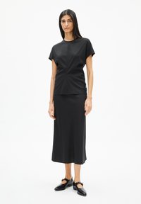 Schwarzes, figurbetontes Oberteil mit kurzen Ärmeln, kombiniert mit einem schwarzen Midikleid. Der Stoff ist glatt und hat einen leichten Glanz. Schwarze Schuhe runden das Outfit ab.