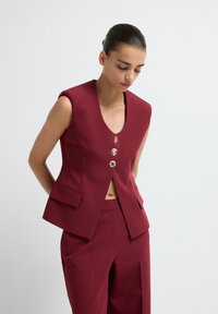 Gilet sur mesure bourgogne avec un col en V, quatre boutons dorés et des poches latérales. Porté avec un pantalon taille haute assorti, texture lisse.