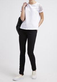Bílý top s síťovanou strukturou a kulatým výstřihem, kombinovaný s černými skinny jeans a bílými teniskami se zlatými akcenty. Batoh přehozený přes rameno.