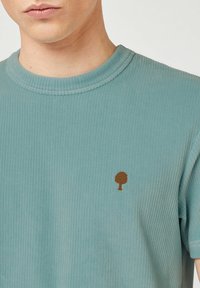 Close-up van een persoon die een lichtblauw ribgebreid T-shirt draagt met een klein bruin boompje-logo op de linkerkant van de borst.