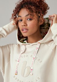 Cremiger Hoodie mit einem blum patterned Innenfutter, mit strukturiertem Logo und gebrandeten Kordeln mit metallischen Akzenten. Weicher Baumwollmischgewebe.