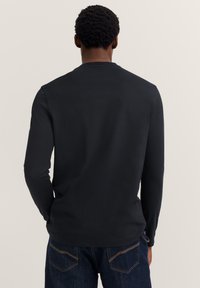 Mann mit dunklem, lockigem Haar, von hinten gesehen, trägt ein schlichtes, schwarzes Langarmshirt und dunkelblaue Jeans vor einem hellen Hintergrund.