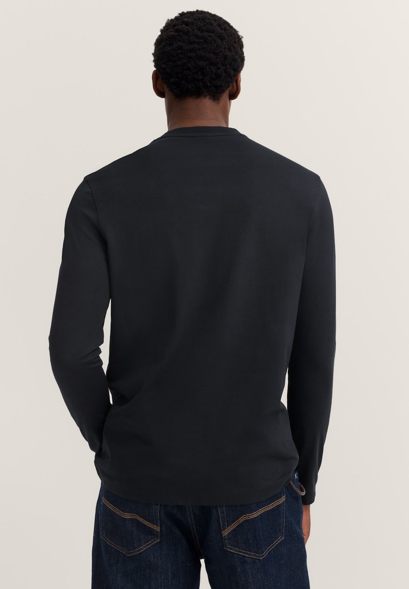 Mann mit dunklem, lockigem Haar, von hinten gesehen, trägt ein schlichtes, schwarzes Langarmshirt und dunkelblaue Jeans vor einem hellen Hintergrund.