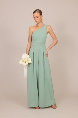 Nő menta zöld egyrészes overálban, egy vállon, fehér kála liliomokat tartva, arany fülbevalókat és szandált viselve, bézs háttér előtt állva.