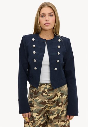Femme portant une veste militaire courte bleu marine avec des boutons argentés sur un haut blanc et un pantalon à motif camouflage.