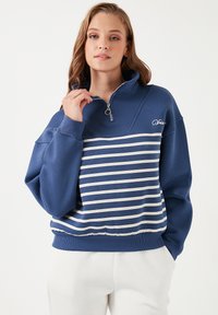 Felpa zip-up blu navy con strisce orizzontali bianche sul torso. Presenta un colletto alto e polsini a coste. Dettaglio del logo semplice sul petto.