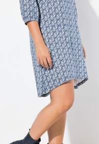 Ein blau-weiß gemustertes Kleid mit Dreiviertelärmeln, das einen geschwungenen Saum und eine bequeme Passform bietet, kombiniert mit dunklen Knöchelstiefeln.