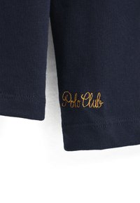 Marinblå stickad pikétröja med ribbad struktur, med guldfärgad kursiv brodyr av "Polo Club" nära fållen.