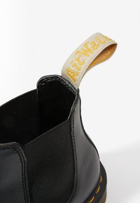 Bottine en cuir noir avec panneau latéral élastique et languette en tissu beige avec texte de marque brodé jaune.
