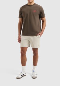 Bruine katoenen T-shirt met rood logo, gecombineerd met lichtbeige shorts. Model draagt witte sneakers met grijze en zwarte accenten. Korte mouwen, ronde hals.