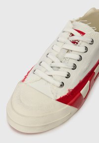 Weißes Canvas-Sneaker mit roten Akzenten, spitz zulaufender Zehenpartie, strukturiertem Sohlenmaterial und sechs Ösen. Das Obermaterial weist fransige Details und kontrastierende Nähte auf.