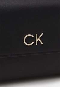 Černá kožená taška s označením "CK" ve zlaté barvě. Hladká textura s jemnými detaily šití, prezentující moderní, minimalistický design.
