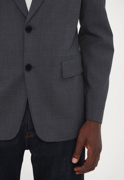 Grauer Blazer aus strukturiertem Stoff, mit zwei schwarzen Knöpfen, einer einzigen Brusttasche und langen Ärmeln, die über dunklen Jeans getragen wird.