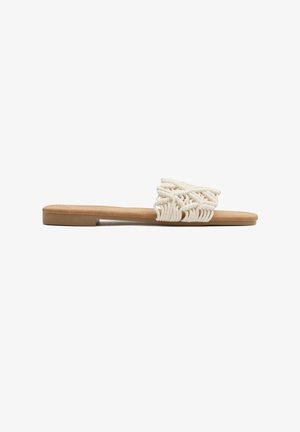 Sandalia plana de color beige con una amplia correa de cuerda crema, intrincadamente anudada en la parte superior, y un tacón bajo.