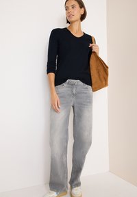 Camicia a maniche lunghe blu navy, jeans chiari larghi e sneakers bianche. Borsa in suede marrone con texture morbida. Design minimalista su uno sfondo neutro.