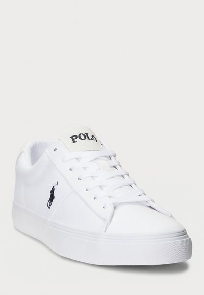 Vita canvas sneakers med platt sula, med en svart broderad logotyp på sidan och texten "POLO" på plösen. Snörning.