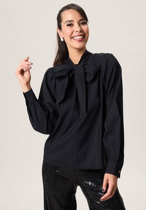 Glimlachende vrouw in een zwarte blouse met lange mouwen en een grote strik bij de hals, gecombineerd met zwarte paillettenbroek, poserend met één hand omhoog.