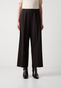 Zwarte wijde broek met plooien aan de voorkant, zachte stof, hoge taille, gecombineerd met zwarte enkellaarzen. De textuur is zacht, de kleur is geheel zwart.