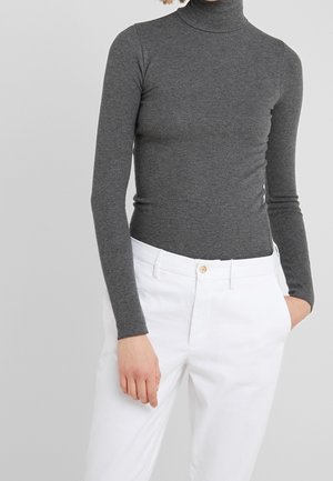 Personne portant un pull à col roulé côtelé gris foncé ajusté et un pantalon blanc taille haute, une main dans la poche, sur un fond uni.