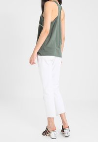 Zielony top bez rękawów z okrągłym dekoltem, zestawiony z białymi spodniami typu crop. Modelka nosi czarne sandały na obcasie z marmurowym wzorem.