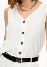 Valkoinen hihaton paita, jossa on V-kaula-aukko ja kuusi mustaa nappia edessä, yhdistettynä mustaan krokotiilikuosisella vyöllä ja kultaisella soljella.