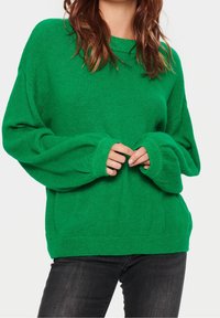 Vihreä ylipitkä villapaita, jossa on teksturoitu neulonta, leveät hihansuut ja pyöreä pääntie. Yhdistetty tummiin farkkuihin kontrastiksi.