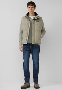 Lichtgrijs puffer-vest over een groene, getextureerde trui, blauwe jeans en bruine laarzen. Het vest heeft een rits- en drukknoopsluiting met zijzakken.