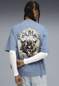 Lyseblå T-shirt med korte ærmer, der har et stort grafisk motiv af en brølende sort panter og ordet "PUMA" i en vintage stil.