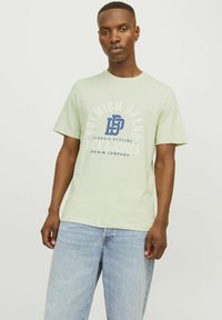 Gaiši zaļa īsām piedurknēm T-krekls ar apaļu kakla izgriezumu, ar zilu grafisku tekstu un logotipu. Valkāts ar gaiši ziliem džinsiem. Gluda auduma tekstūra.