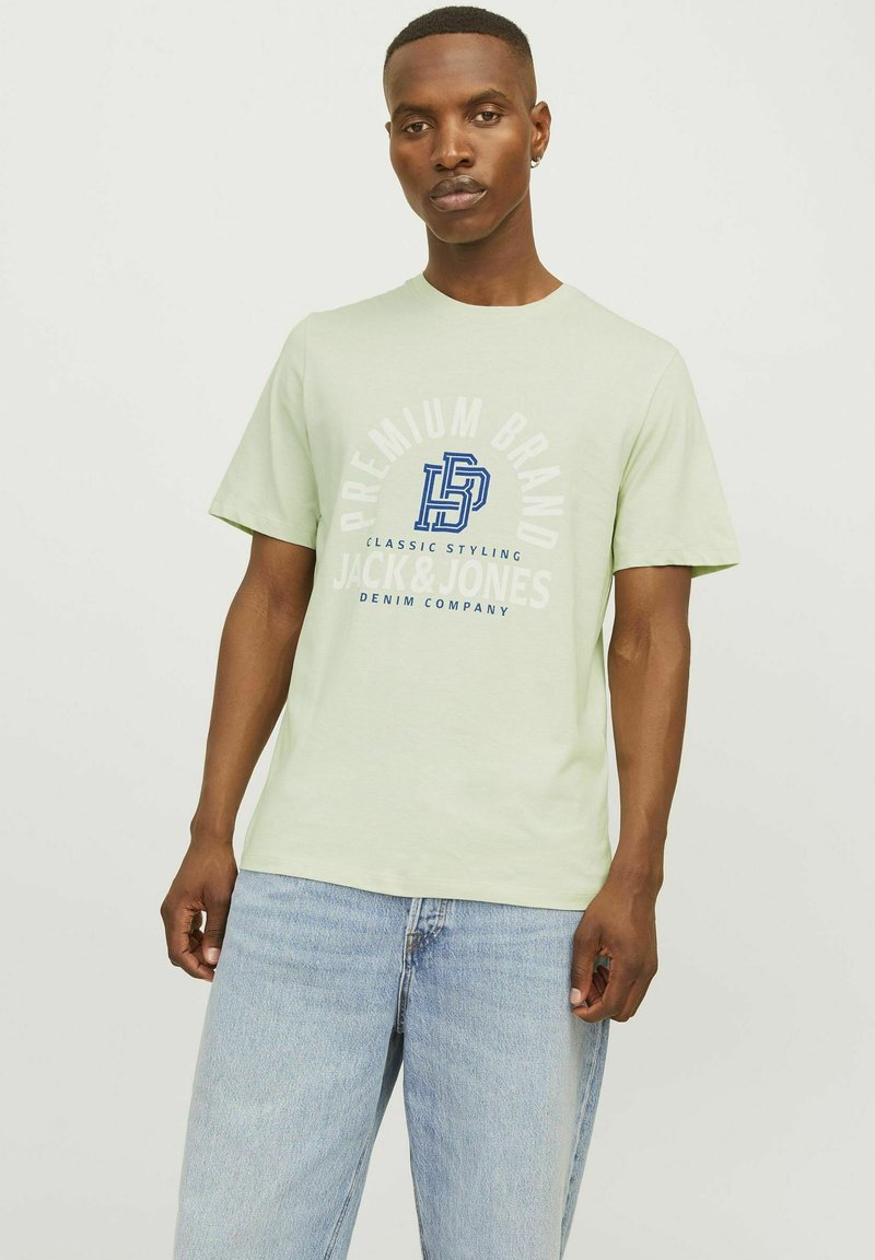 Gaiši zaļa īsām piedurknēm T-krekls ar apaļu kakla izgriezumu, ar zilu grafisku tekstu un logotipu. Valkāts ar gaiši ziliem džinsiem. Gluda auduma tekstūra.