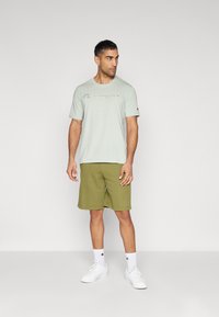 Hellgrünes Baumwoll-T-Shirt mit "Champion"-Logo, kombiniert mit olivgrünen Shorts. Weiße Socken und Sneakers runden das Outfit ab.