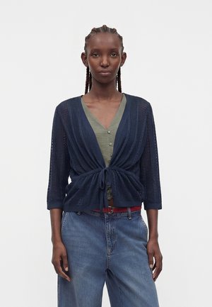 Vrouw met ingevlochten haar die een donkerblauwe losse cardigan draagt over een groene top met knopen en een hooggesloten blauwe spijkerbroek met een rode riem.