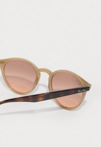 Γυαλιά ηλίου Ray-Ban με ελαφρύ μπεζ πλαστικό σκελετό και gradient ροζ φακούς. Οι γέφυρες με σχέδιο καφέ προσφέρουν ασημένια λεπτομέρεια λογότυπου.