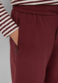Hand in pocket van bordeauxkleurige, gestructureerde broek met rits en haakje, gedragen met een lange mouw bordeauxkleurig en wit gestreept shirt.