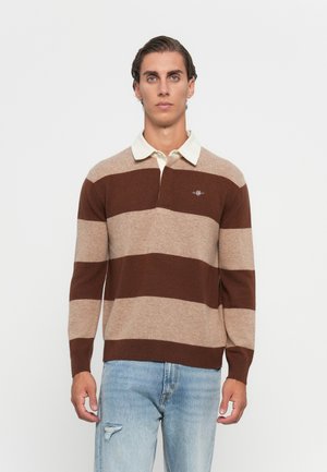Man die recht vooruit kijkt, gekleed in een bruin en beige breedgestreept rugbyshirt met een witte kraag en lichtblauwe versleten spijkerbroek.
