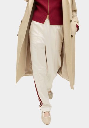 Beiger langer Mantel über rotem Reißverschluss-Pullover und weißen Hosen mit rotem Seitenstreifen, kombiniert mit beigen Slip-On-Schuhen, geht vorwärts.