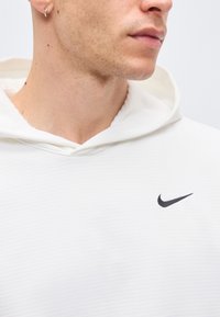 Κοντινό πλάνο ενός ατόμου που φοράει λευκό φούτερ Nike με κουκούλα, εμφανίζοντας τον λαιμό, το κάτω μέρος του προσώπου και το μαύρο λογότυπο Nike στη λεκάνη.