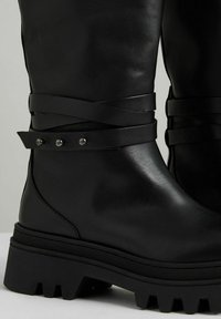 Svarta läderknähöga stövlar med korsade remmar och metallnitar vid vristen, med en kraftig sula för bra grepp.