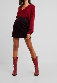 Femme portant un pull col en V bordeaux, une mini-jupe en velours côtelé violet foncé et des bottes de cow-boy rouges, debout devant un fond uni.