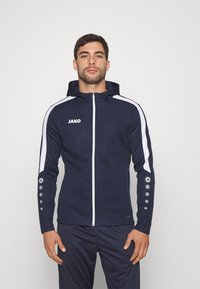 Marineblauwe sportjas met een slim fit ontwerp, witte strepen op de schouders en knopenaccenten op de mouwen. Voorzien van een capuchon en een ritssluiting aan de voorkant.